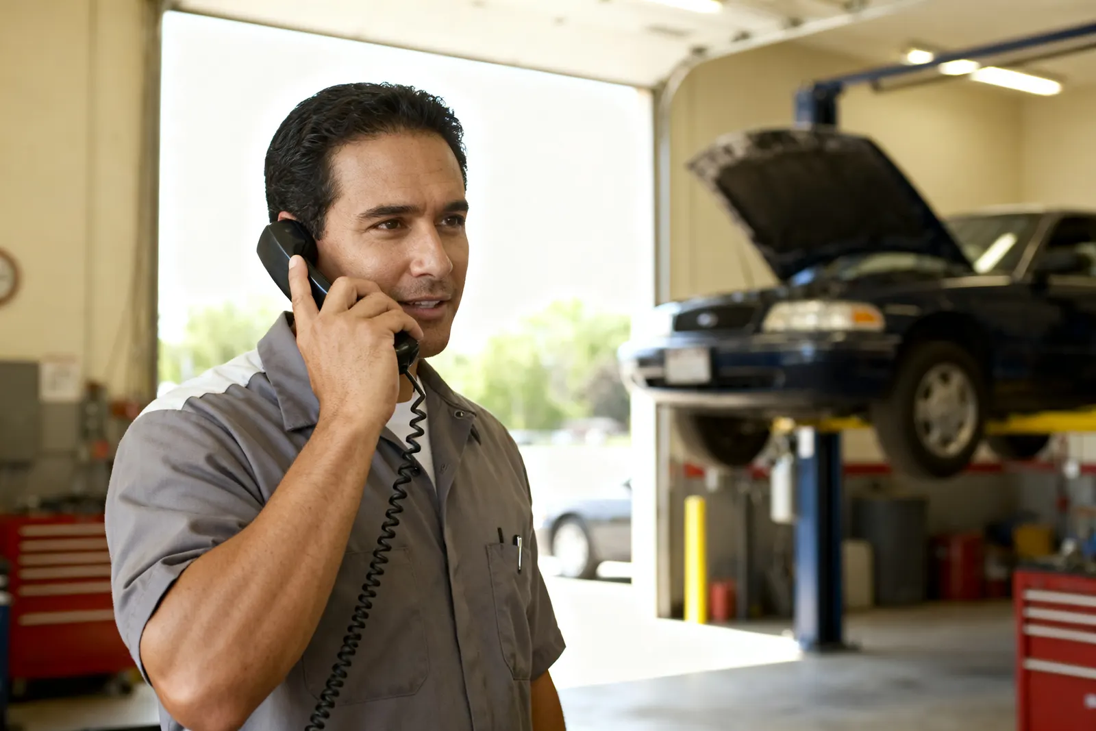 Mechanic taking a customer call in the shop with a car on the lift behind him — Rinqly's AI automotive answering service answers every call 24/7